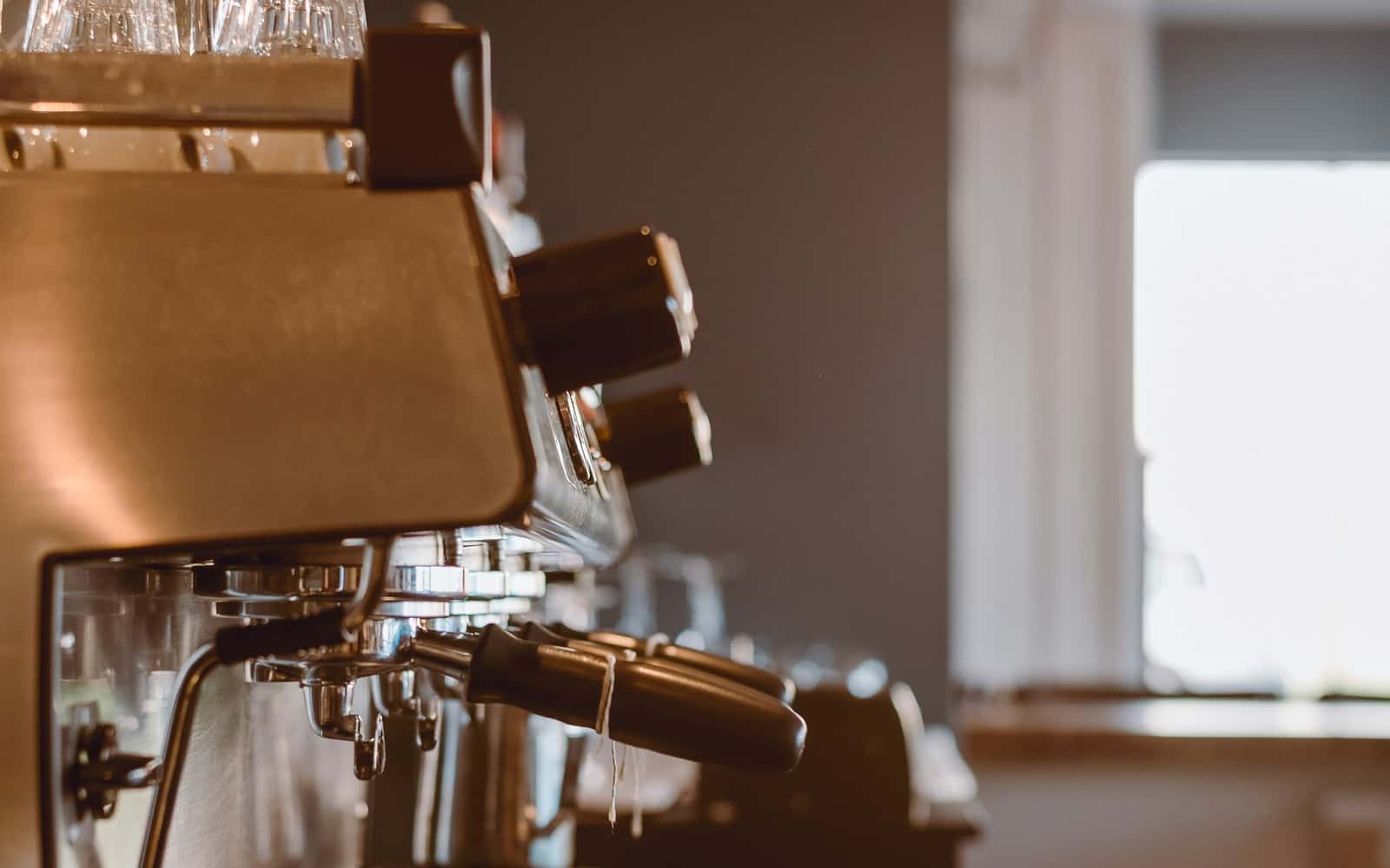 Der Brühkopf einer Espressomaschine, von der Seite beleuchtet, mit Gläsern auf der Tassenablage