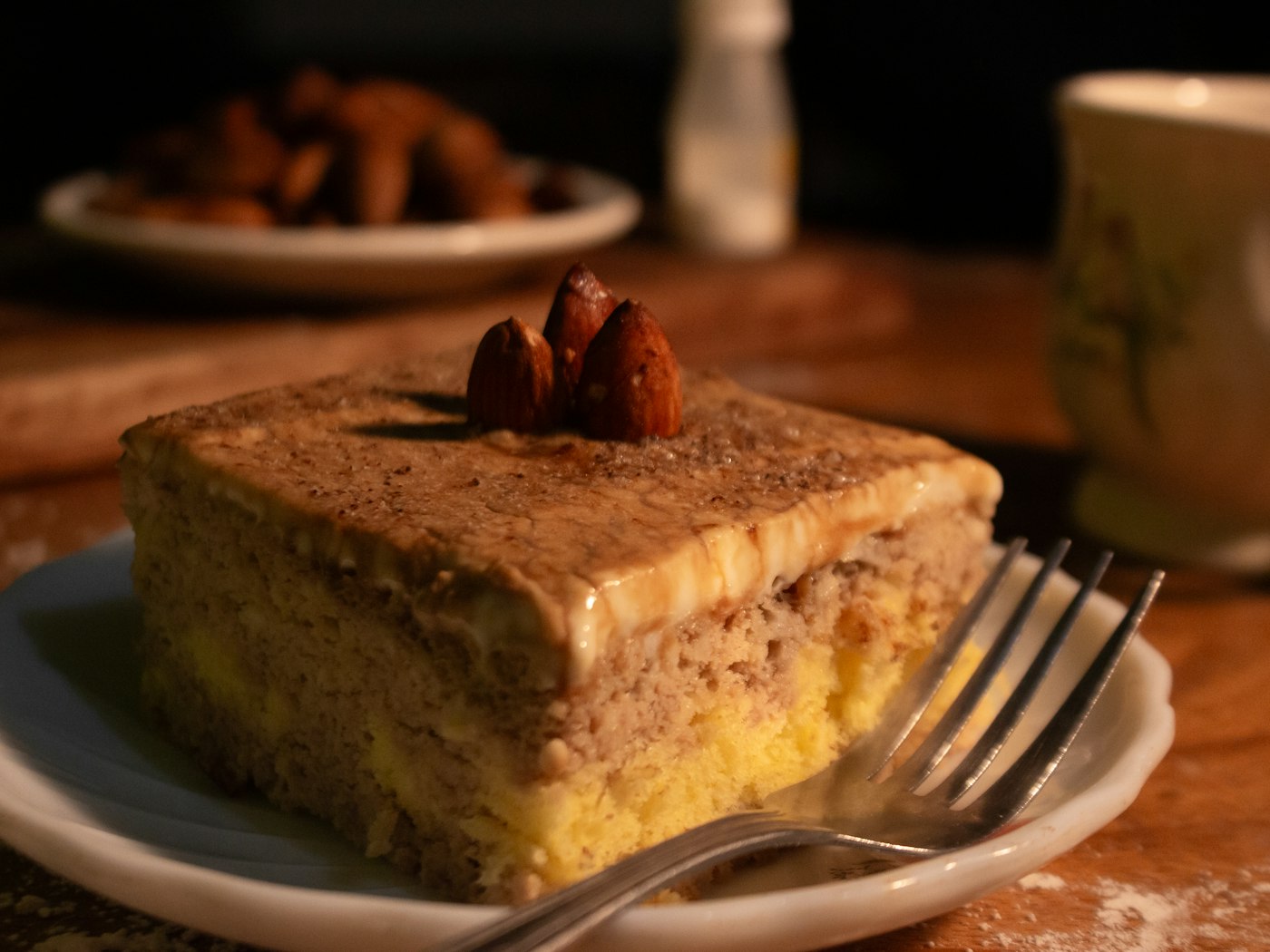 Ein eckiges Stück Kuchen mit Cremeschicht und ganzen Mandeln obenauf, auf einem Teller im warmen Halbdunkel