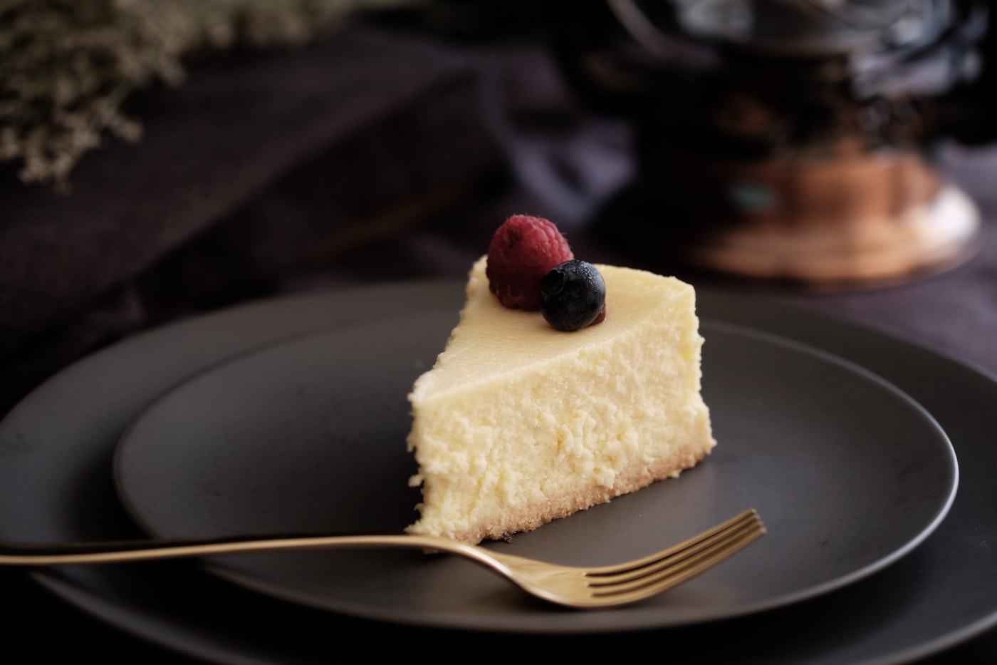 Ein hohes Stück Käsekuchen auf einem dunkelgrauen Teller, daneben eine Gabel