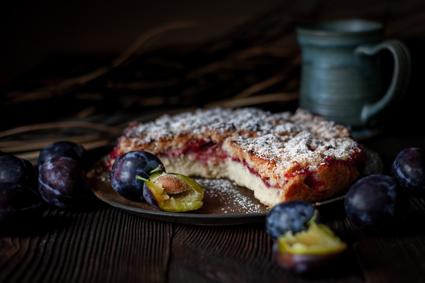 Ein angeschnittener Pflaumenkuchen mit Streuseln und Puderzucker, ringsum ganze und halbierte Pflaumen auf dunklem Holz